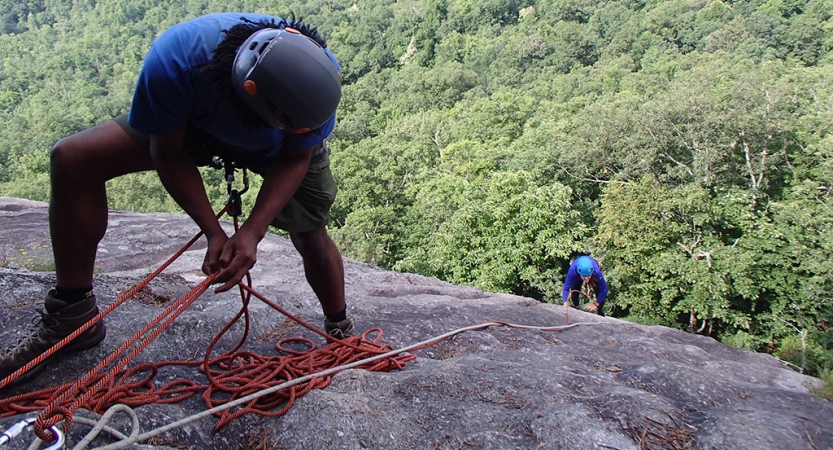 In the foreground, one person wearing safety gear is secured by ropes as they look down a rocky slope to another person, also wearing safety gear and secured by ropes, who is making their way up.